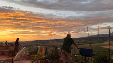 Duas pessoas e um cachorro branco estão em um terraço, contemplando um pôr do sol de tirar o fôlego sobre colinas ondulantes, com nuvens vibrantes e toalhas coloridas penduradas nas proximidades.