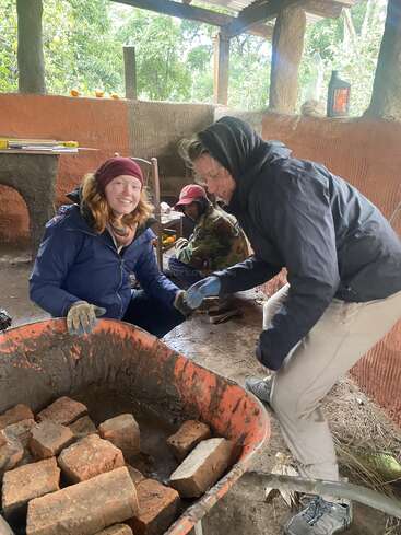 Drei Personen arbeiten zusammen in einem Haus und hantieren mit Ziegeln und Baumaterialien. Sie sind warm angezogen, lächeln und scheinen an einem Bau- oder Gemeinschaftsprojekt beteiligt zu sein.