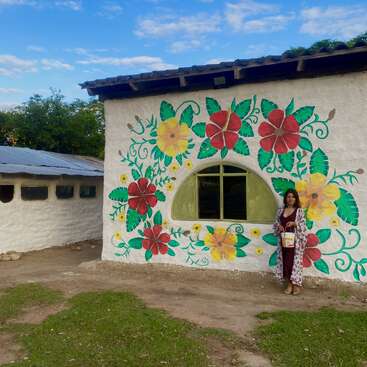 Una mujer está de pie junto a un edificio blanco decorado con un vibrante mural floral rojo y amarillo, bajo un cielo azul con nubes dispersas, rodeada de vegetación y belleza natural.