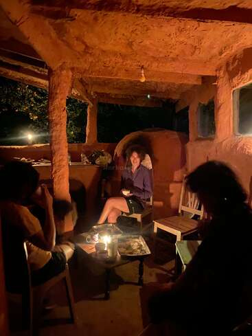 Three people relax in a rustic, earthy room lit by candlelight. The cozy ambiance suggests a warm, intimate evening with friends, conversation, and shared snacks.