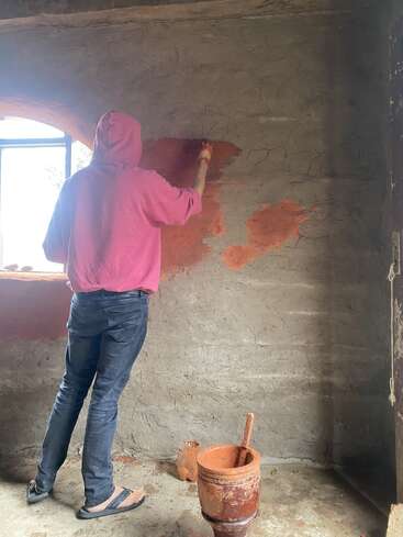 A person in a pink hoodie paints a textured wall with reddish-brown paint. Light enters through a window. A paint bucket and brush rest nearby.