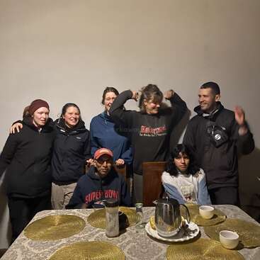 Seven people, five standing and two sitting, gather around a dining table with placemats and cups. Some are laughing and making funny gestures, enjoying themselves.