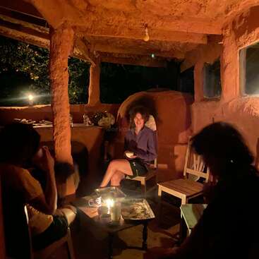 Three people relax in a rustic, earthy room lit by candlelight. The cozy ambiance suggests a warm, intimate evening with friends, conversation, and shared snacks.