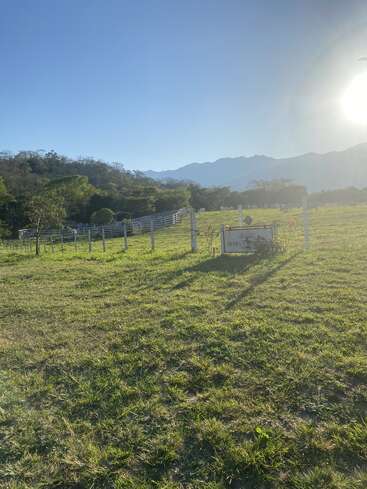 Dieses Bild zeigt eine sonnenbeschienene Wiese mit einem weißen Zaun, einige Bäume, Berge im Hintergrund, einen klaren blauen Himmel und ein Schild im Vordergrund.