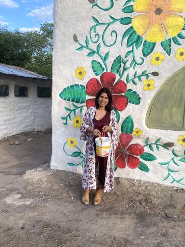 Eine Frau steht lächelnd vor einer bunt bemalten Wand mit bunten Blumen. Sie hält einen Farbeimer in der Hand, trägt Stiefel und ein geblümtes Gewand. Draußen, ein sonniger Tag.