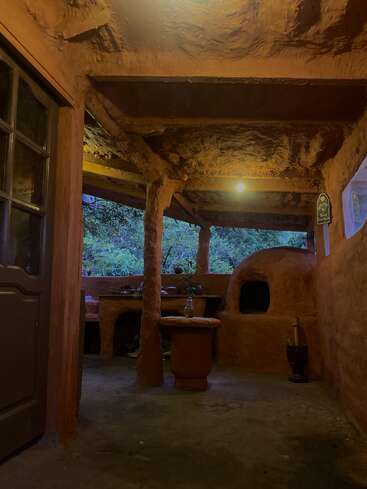 This cozy rustic kitchen features earthen walls, a traditional clay oven, wooden furniture, warm lighting, and lush greenery outside the open window, creating a peaceful atmosphere.