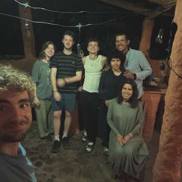 A group of six people pose happily together at night, under string lights and rustic pillars. One person sits in front while others stand behind, smiling warmly.