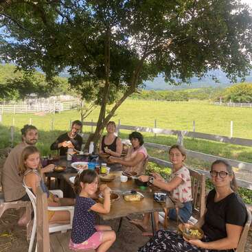 Eine Gruppe von Leuten, darunter Erwachsene und Kinder, sitzt draußen unter einem großen Baum an einem Holztisch und isst was. Um sie herum gibt's grüne Wiesen und Zäune.