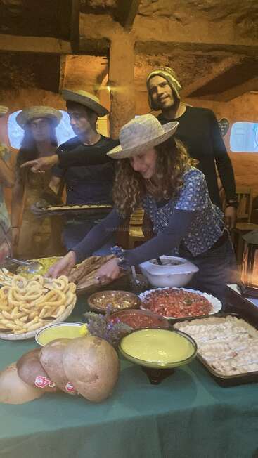 Um grupo animado, usando chapéus, se reúne em torno de uma mesa repleta de diversas comidas tradicionais, pães e molhos, dentro de um ambiente rústico e bem iluminado.