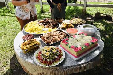 A round outdoor table is filled with a variety of colorful dishes, cake with pink roses, salad, rice, meat, bananas, and sauces, in sunlight.