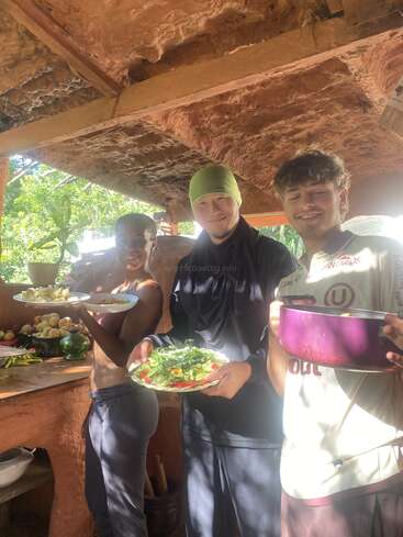 Three smiling people stand in a rustic outdoor kitchen, holding plates of fresh food and a pot, surrounded by natural light, fruit, and earthy, wooden surroundings.