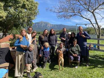 A group of people and two dogs gather outdoors on a sunny day, smiling together. Trees, grass, mountains, and blue sky provide a beautiful natural backdrop.