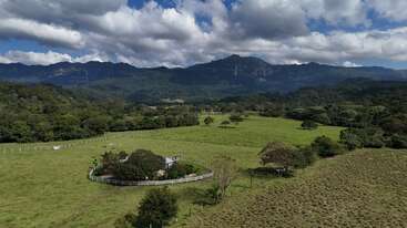 Uma paisagem rural serena com campos verdes abertos, árvores espalhadas, um pequeno cercado, florestas exuberantes e montanhas majestosas sob um céu azul brilhante e parcialmente nublado.