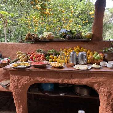 Uma cozinha rústica ao ar livre exibe um banquete colorido de frutas frescas, legumes, jogos de chá e pratos tradicionais. Uma vegetação exuberante e laranjeiras cercam esse ambiente aconchegante.