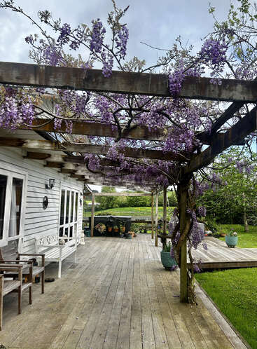 Une terrasse en bois confortable avec des meubles d'extérieur blancs, couverte par une pergola ornée de glycines violettes en fleurs. Des plantes en pot et de la verdure complètent ce cadre paisible.