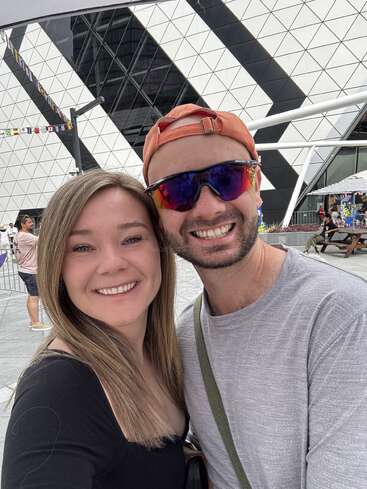 Das Bild zeigt ein Selfie von einem Mann und einer Frau, die vor einem modernen Gebäude mit geometrischem Muster stehen, das wahrscheinlich tagsüber aufgenommen wurde.