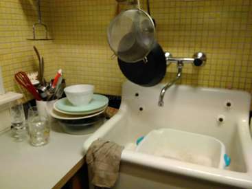 A imagem mostra uma pia de cozinha com uma bacia de porcelana branca, uma panela pendurada e uma pilha de pratos sobre o balcão, em uma parede de azulejos amarelos.