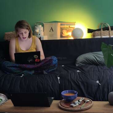 Uma mulher senta-se de pernas cruzadas em um sofá escuro, usando um laptop. Uma lâmpada quente ilumina os livros atrás dela. Uma mesa contém sopa, pão e outro laptop. Ambiente aconchegante.