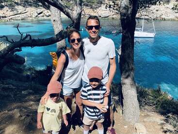 La imagen muestra a una familia de cuatro miembros posando para una foto en una orilla rocosa, con una serena masa de agua y una pequeña embarcación al fondo.