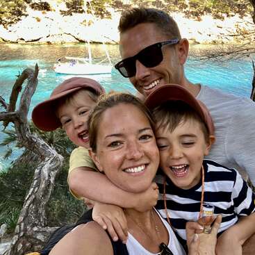 La imagen muestra a una familia de cuatro miembros posando para un selfie frente a una serena masa de agua, con un barco visible al fondo.