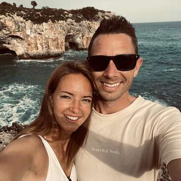 Das Bild zeigt ein Paar, das ein Selfie vor einer felsigen Küste macht. Die Frau auf der linken und der Mann auf der rechten Seite tragen beide weiße Oberteile.