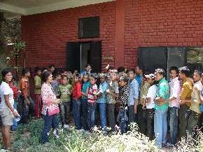A imagem mostra um grupo de aproximadamente 30 pessoas, a maioria crianças, reunidas do lado de fora de um prédio de tijolos vermelhos. Elas estão em pé em uma área gramada.