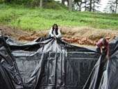 A imagem mostra dois indivíduos trabalhando em uma grande lona preta que cobre um buraco no chão, situado em uma área gramada com árvores ao fundo.