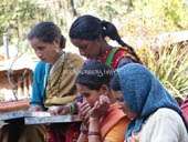 A imagem mostra quatro mulheres reunidas em torno de uma mesa, sendo que uma delas usa um lenço de cabeça azul e a outra um sári amarelo. O fundo está embaçado.