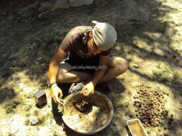 A imagem mostra um homem sentado no chão, cercado por uma coleção de objetos, incluindo uma tigela, um recipiente e uma pilha de pequenas bolas, em um ambiente natural.
