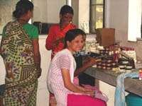 A imagem mostra três mulheres em uma sala, uma delas sentada em uma mesa, cercada por vários itens, enquanto as outras duas estão próximas, envolvidas em uma atividade.