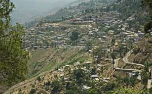 A imagem retrata uma pequena cidade situada em um vale, com uma estrada sinuosa que leva à cidade e uma encosta coberta de árvores e edifícios.