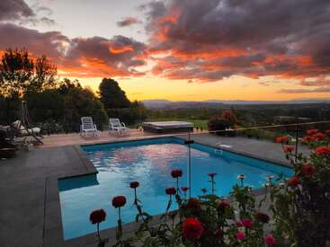 Uma cena serena à beira da piscina ao pôr do sol, com nuvens laranja vibrantes refletindo na água, cercadas por flores, espreguiçadeiras e colinas distantes sob um céu dramático.