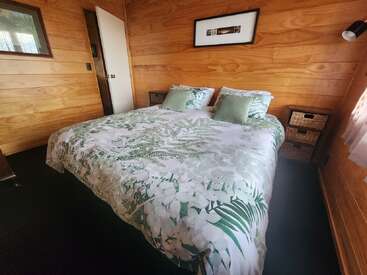 Este quarto aconchegante tem paredes de madeira, uma cama grande com roupa de cama com tema botânico, duas mesas de cabeceira pequenas com gavetas de tecido, iluminação suave e uma janela com cortinas transparentes.