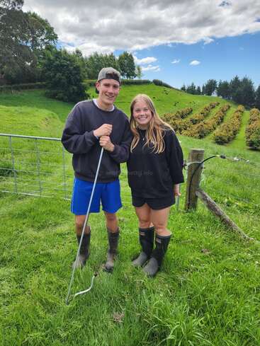 Dois jovens sorriem na grama verde exuberante, usando botas e roupas casuais. Atrás deles, há uma paisagem agrícola montanhosa e cênica com árvores e arbustos.