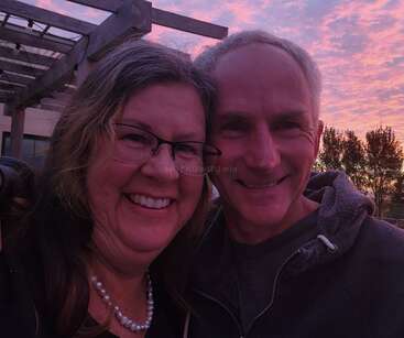 Um casal sorridente tira uma selfie ao ar livre no pôr do sol, apreciando o céu colorido em tons de rosa e roxo. Você pode ver as árvores e a estrutura de uma pérgola ao fundo.
