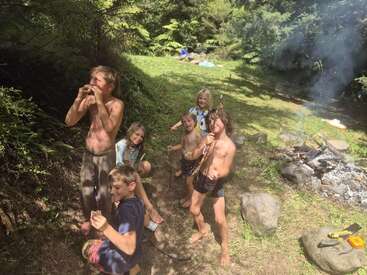 Six children enjoy an outdoor adventure in a lush, green forest, roasting food over a campfire, laughing and bonding together under the bright sunlight.