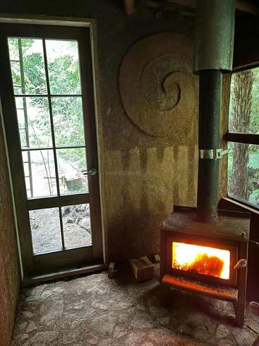 This cozy room features a wood-burning stove radiating warmth, a glass door revealing lush greenery outside, textured walls with a spiral design, and rustic stone flooring.