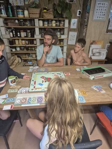 A group of people, including children and an adult, are playing Monopoly around a wooden table in a cozy, rustic kitchen filled with jars and plants.