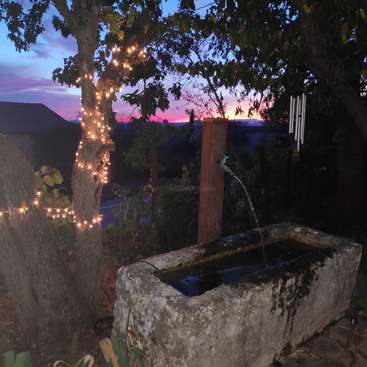 The image depicts a serene outdoor setting at dusk, featuring a stone fountain, a tree adorned with lights, and a breathtaking sunset in the background.