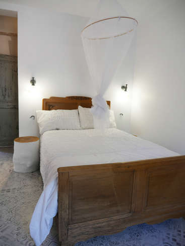 The image depicts a serene bedroom featuring a wooden bed frame, white bedding, and a mosquito net, set against a crisp white wall and patterned floor.