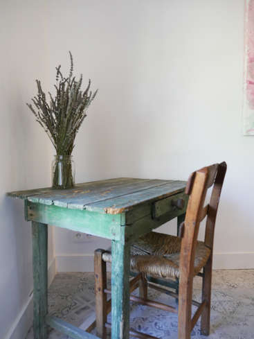 The image depicts a rustic wooden table with a drawer, accompanied by a wooden chair and a vase of dried flowers, set against a white wall and tiled floor.
