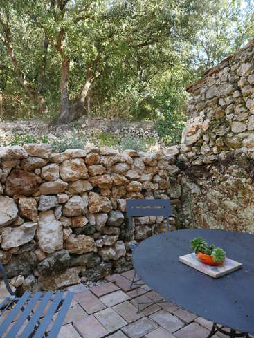 The image depicts a serene outdoor patio area, featuring a stone wall, a round table with a plant, and a metal chair, surrounded by lush trees and foliage.