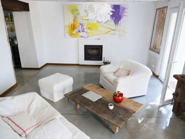 The image depicts a modern living room with white walls, furniture, and a large abstract painting above a fireplace, featuring a wooden coffee table and a doorway to a kitchen.