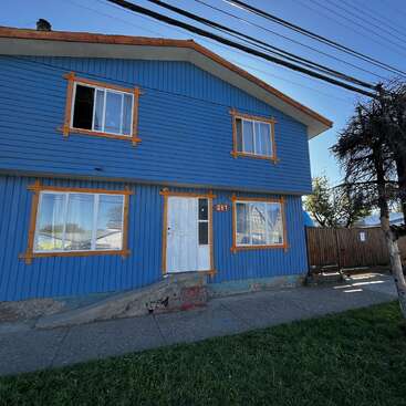 This image shows a blue two-story house with orange window trims, a white door, sloped sidewalk, grassy area, and overhead power lines beneath a clear sky.