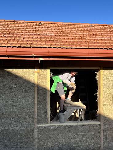 A person stands on a wooden platform inside a window frame of a straw-bale house under construction, with a tiled roof and clear blue sky above.