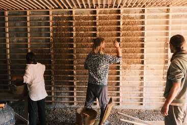 Three people are working together, applying a natural material to a wooden lattice wall framework, possibly constructing an eco-friendly or sustainable building using traditional techniques.