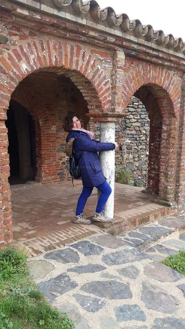 The image depicts a woman posing in front of a brick archway, wearing a blue coat and pants, with a backpack and standing on a stone pathway.