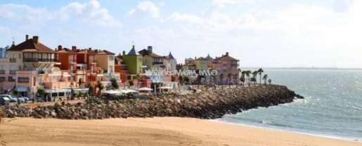 The image depicts a coastal town with a sandy beach, a rocky shoreline, and a row of buildings, set against a cloudy sky.