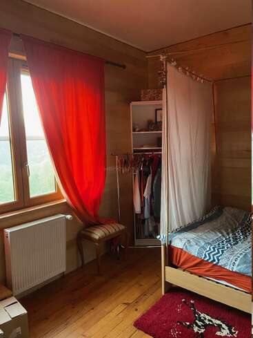 Ein gemütliches Schlafzimmer mit Holzwänden, roten Vorhängen, einem Fenster, einem kleinen Bett mit Baldachin, einem offenen Kleiderschrank, einer Bank, einem Heizkörper und einem roten Teppich mit Muster.