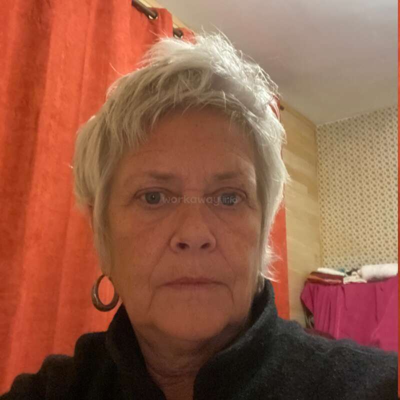 Eine Frau mit kurzen grauen Haaren und Creolen posiert drinnen für ein Selfie; im Hintergrund des Raums sind rote Vorhänge und gemusterte Tapeten zu sehen.
